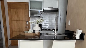 A modern kitchen with a central island, white chairs, stainless steel appliances, and a wooden door. - Modern Ocean View 1BR in La Esperilla, Pool, Gym (Santo Domingo)