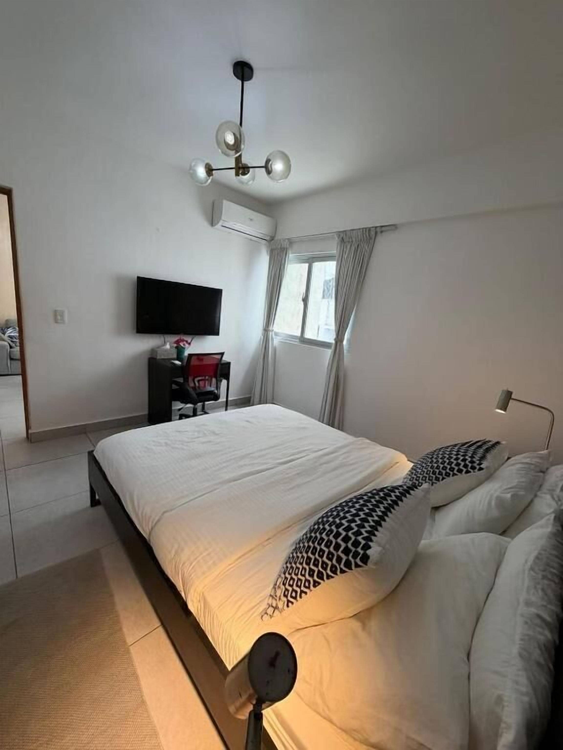 A bedroom with a bed, a flat-screen TV, a desk with a chair, and a window with curtains.