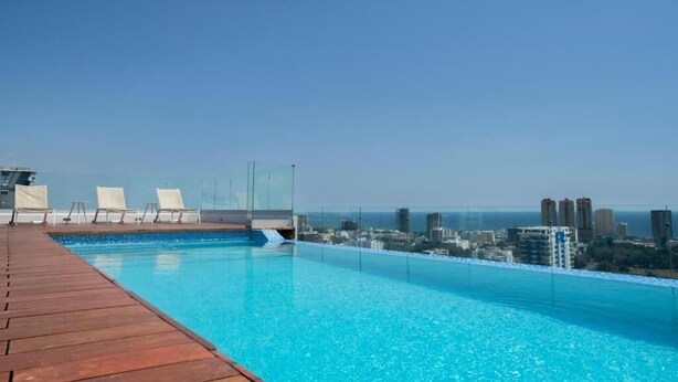 A rooftop infinity pool with a wooden deck, lounge chairs, and a cityscape view. - Modern Ocean View 1BR in La Esperilla, Pool, Gym (Santo Domingo)