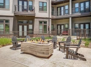 A modern outdoor seating area with a fire pit, chairs, and a building with balconies in the background.