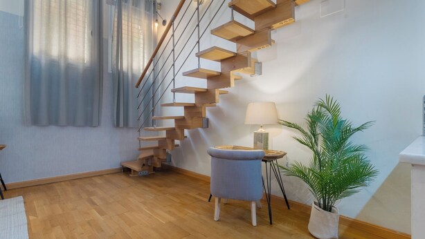 A modern staircase with wooden steps and metal railings, a small round table with a lamp, a potted plant, and a window with curtains. - Exclusive loft in the center of Seville (Sevilla)