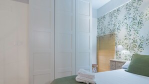 A bedroom with a bed, a nightstand, a lamp, a window with blinds, and a wall with floral wallpaper. - Exclusive loft in the center of Seville (Sevilla)