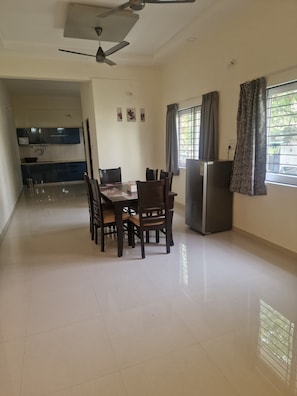 Una habitación amplia con mesa de comedor, sillas, ventilador de techo, refrigerador y ventana con cortinas.
