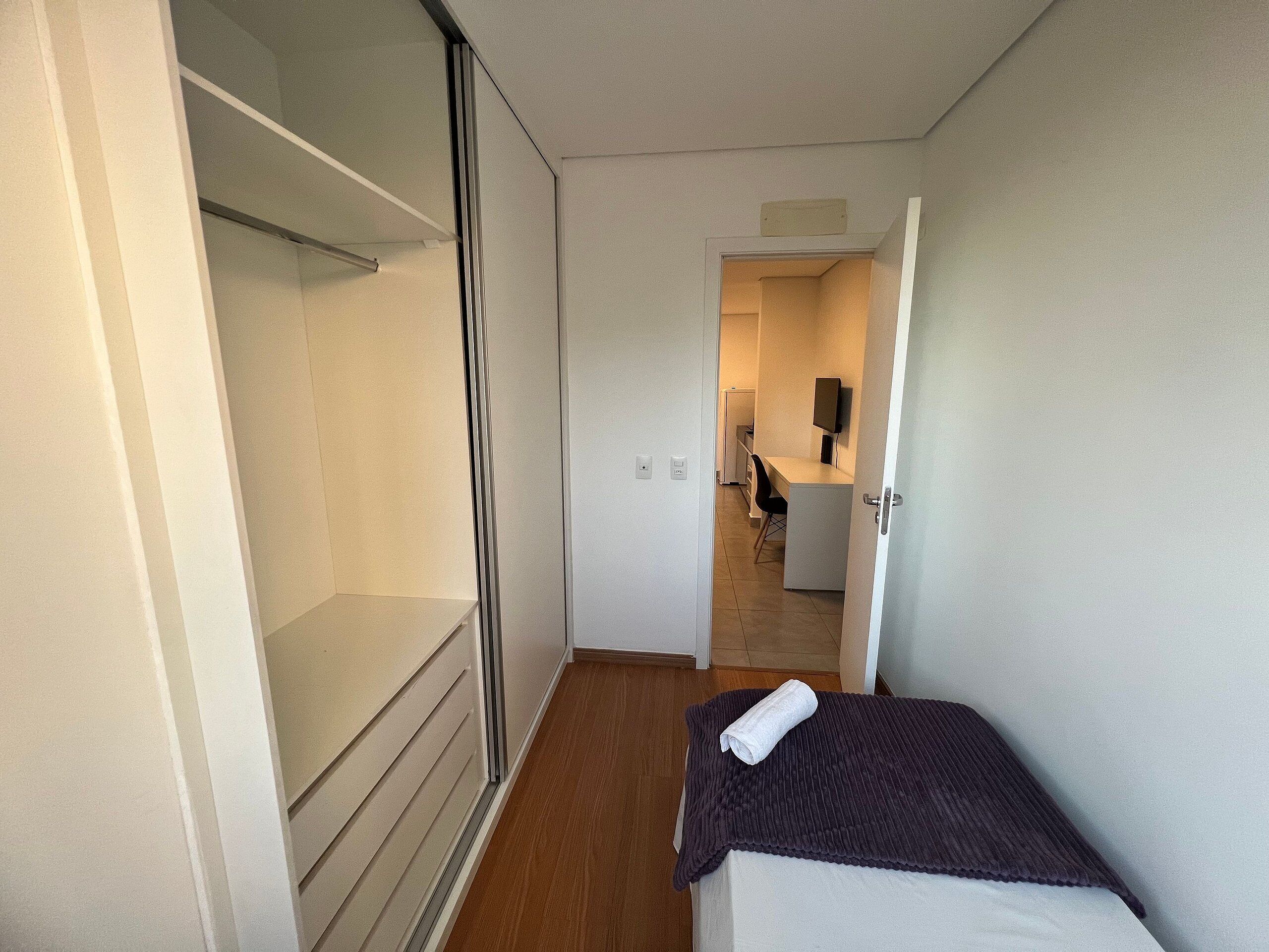 Un pasillo estrecho con una cama, un armario y una kitchenette al fondo.