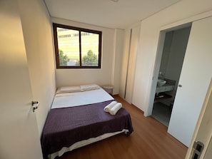 A bedroom with a bed, a window, a small table, and a bathroom with a sink.