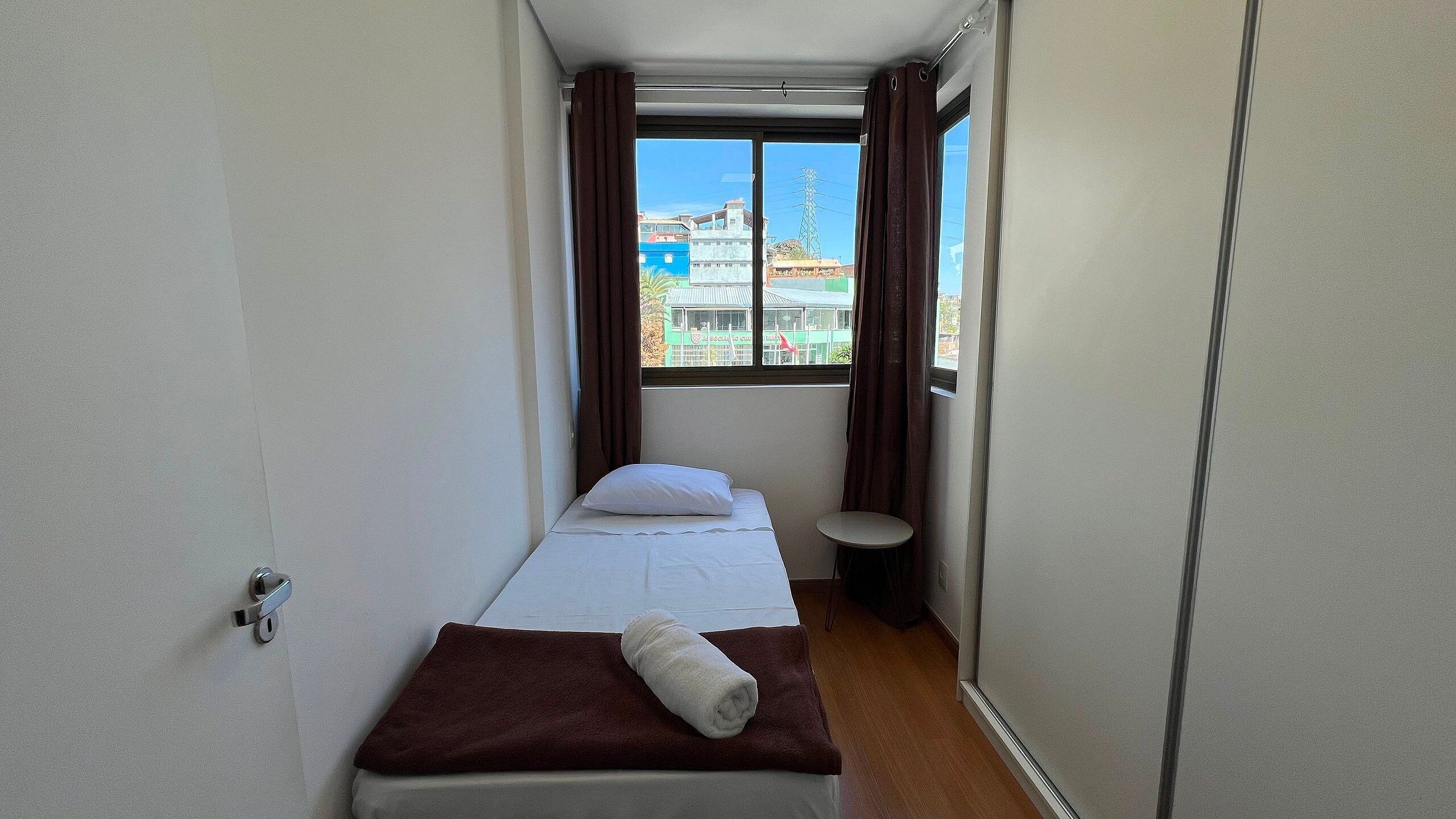 Ein schmales Hotelzimmer mit einem Einzelbett, einem kleinen Tisch und einem fenster mit Vorhängen.