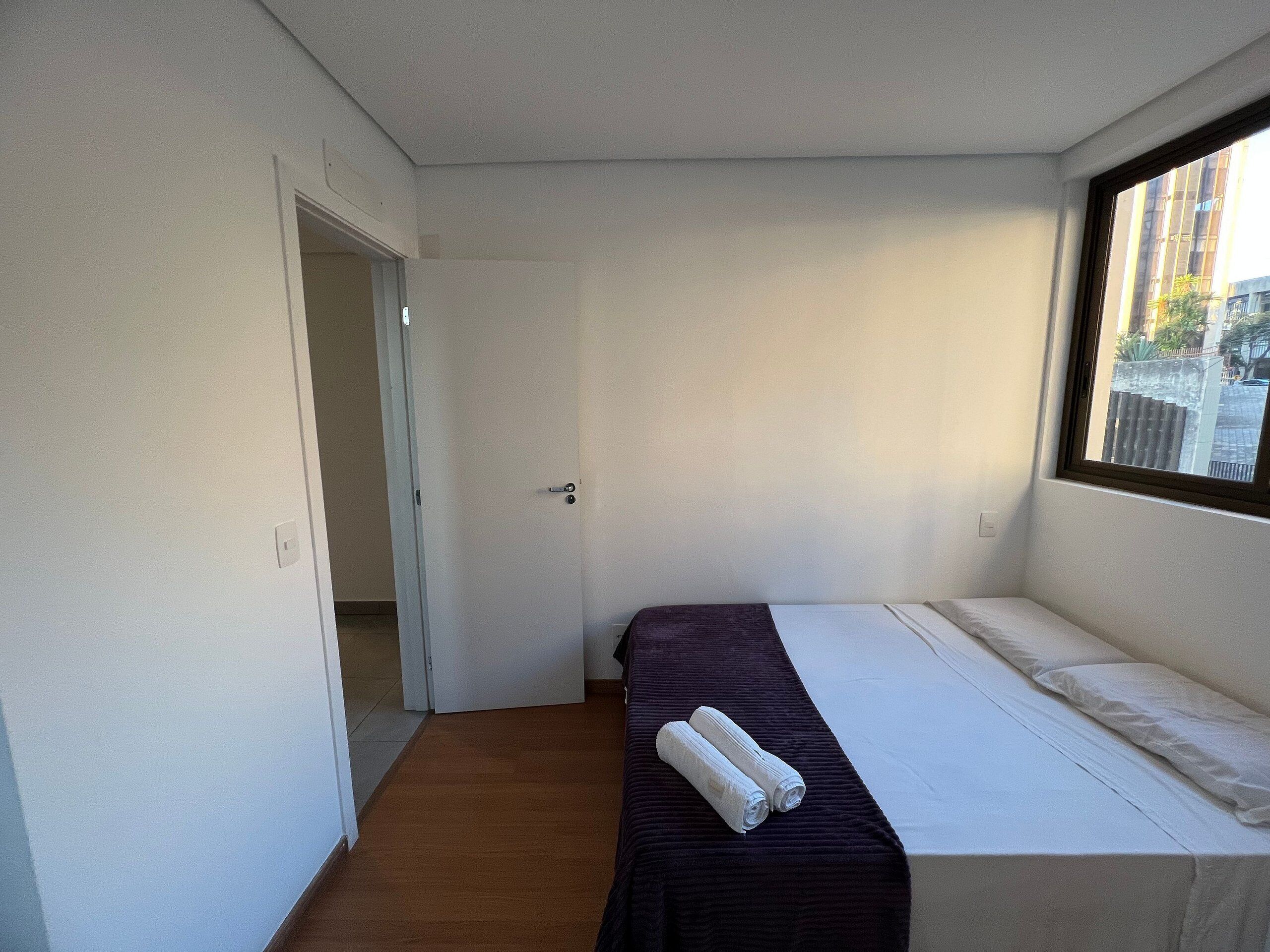 Quarto pequeno com uma cama de solteiro, porta de acesso a outro espaço, janela com vista para prédios e cama com colcha escura e duas toalhas enroladas.
