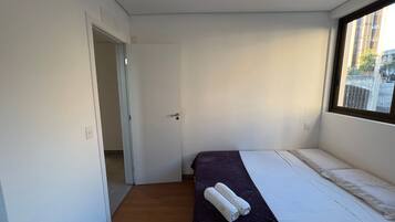 A small, single-bed room with a door leading to another room, a window with a view of buildings, and a bed with a dark-colored bedspread and two rolled-up towels.
