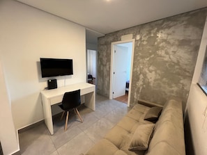Une pièce avec un bureau blanc, une chaise noire, un canapé beige et un téléviseur.