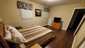 Un dormitorio con una cama, una mesita de noche de madera, un mueble para televisión y una puerta que da a otra habitación.