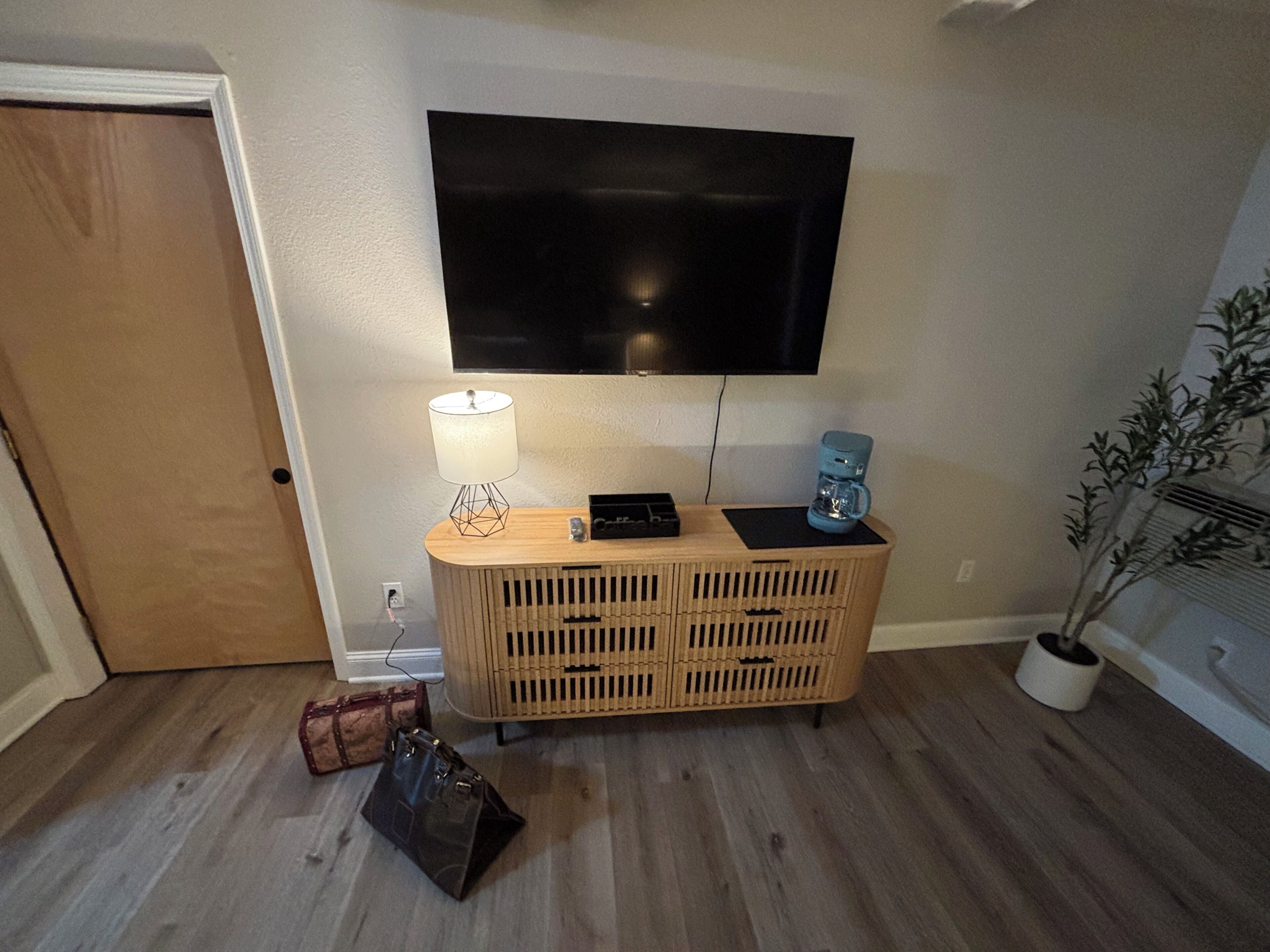 A room with a wooden cabinet, a flat-screen TV mounted on the wall, a lamp on the cabinet, a coffee maker, a suitcase, and a potted plant.