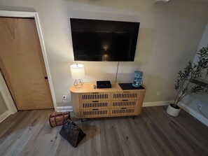 Una habitación con un armario de madera, un televisor de pantalla plana montado en la pared, una lámpara sobre el armario, una cafetera, una maleta y una planta en maceta.
