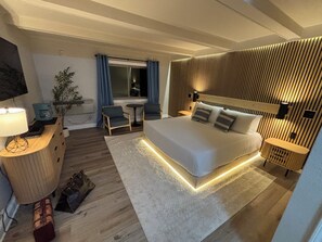 Habitación de hotel moderna con una cama grande, mesitas de noche, un escritorio, una silla, una pequeña mesa y un televisor.