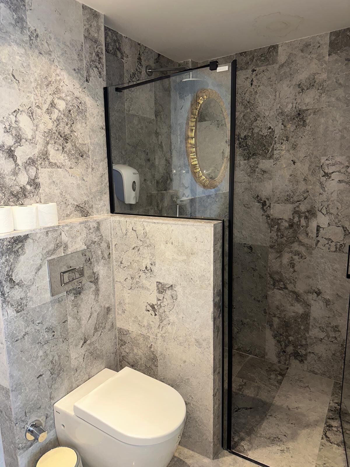 Un baño con inodoro, ducha con mampara de vidrio y pared de mármol.