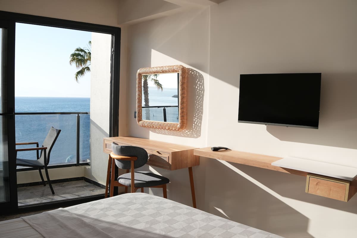 A bedroom with a bed, a desk with a chair, a TV, and a balcony with a view of the sea.