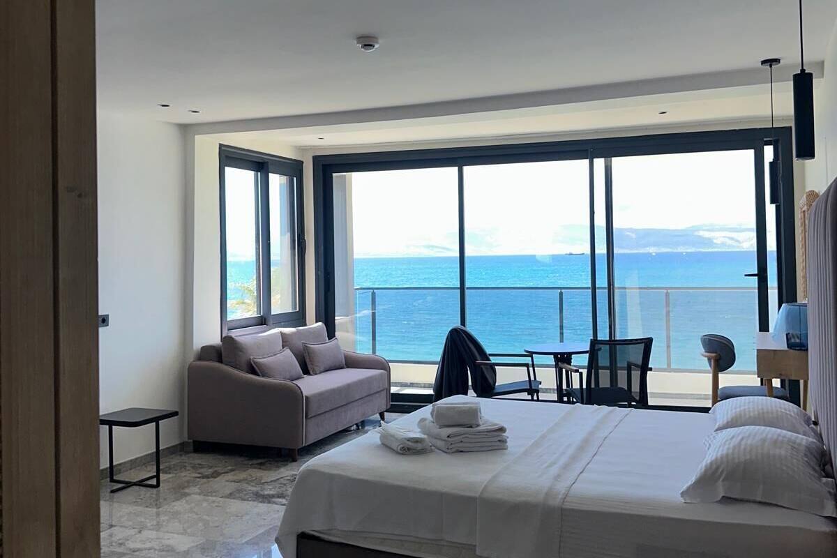 A modern hotel room with a large bed, a sofa, a dining area, and a view of the sea.