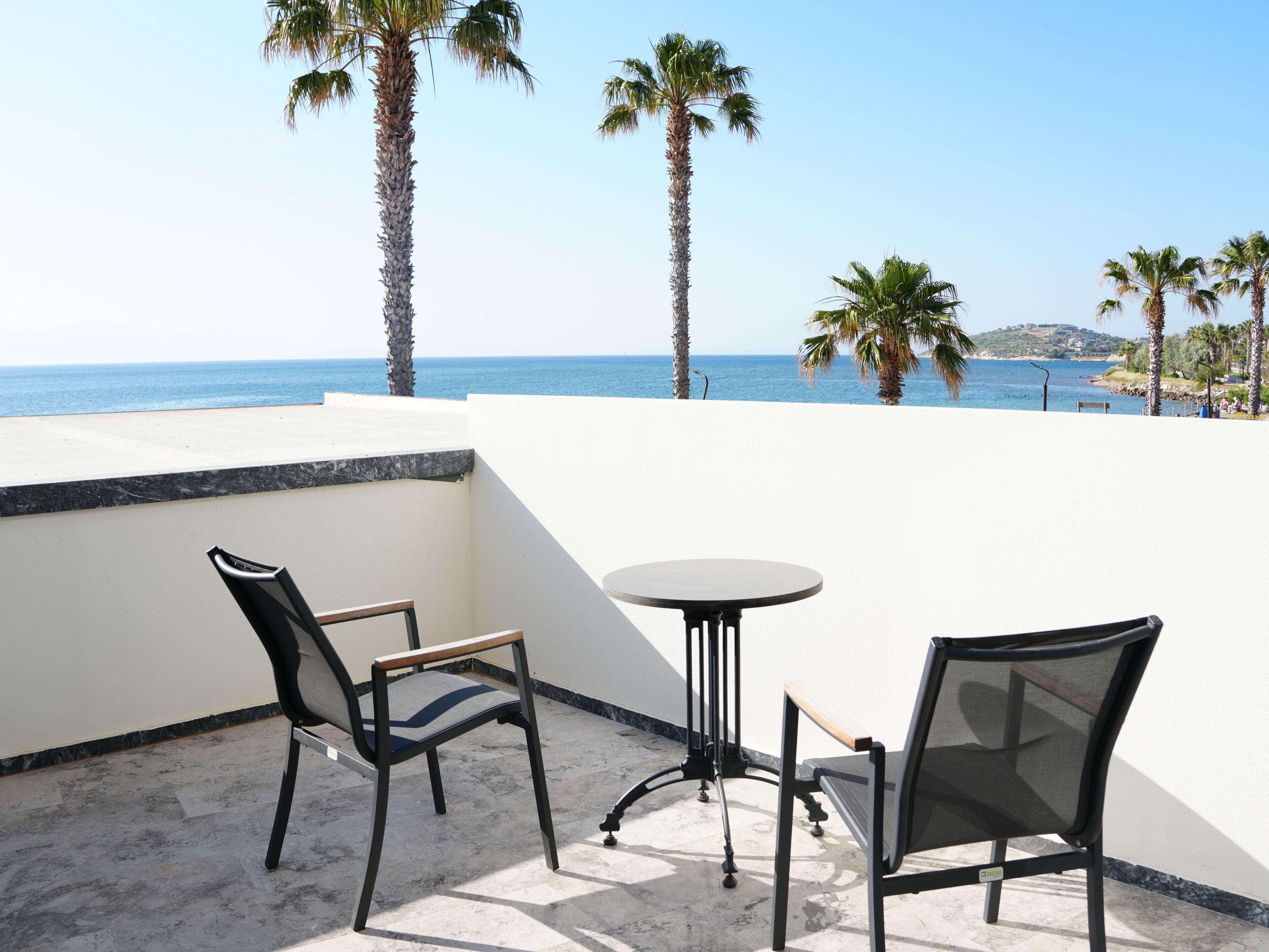 Terraço com duas cadeiras e uma mesa, com vista para uma praia com palmeiras e o mar.