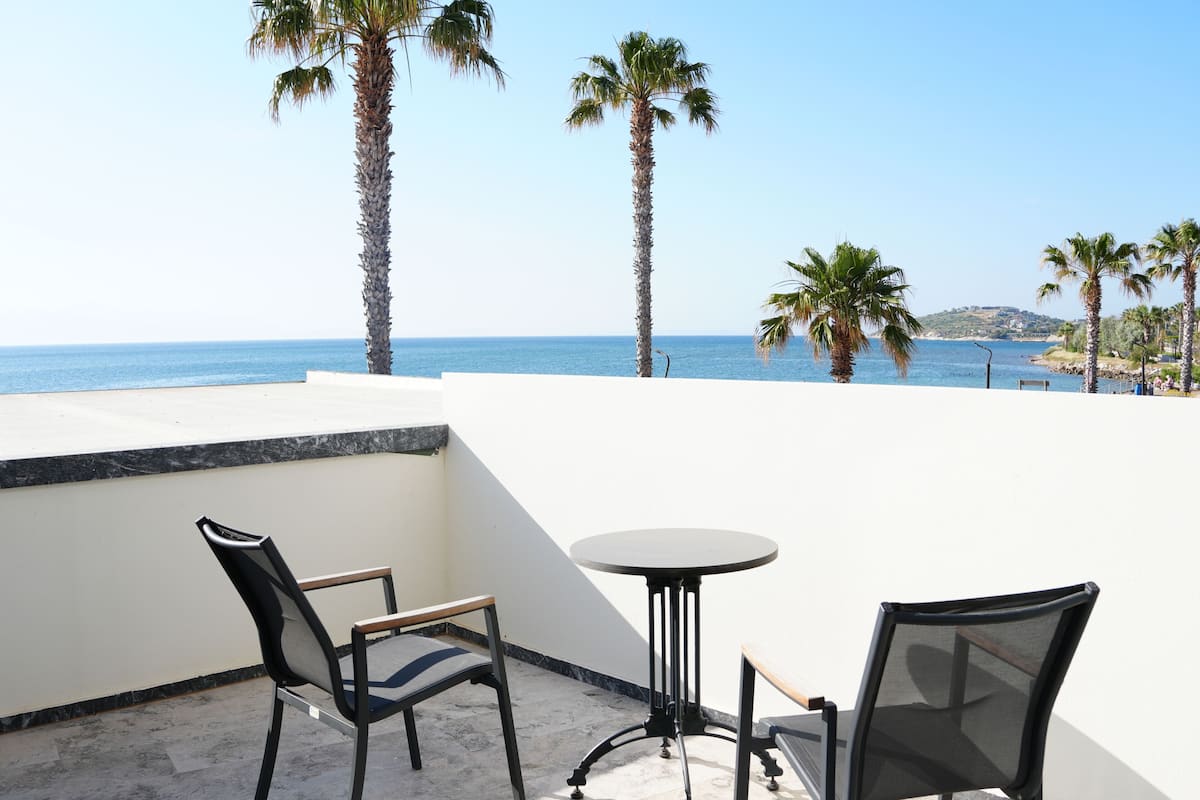 A terrace with two chairs and a table, overlooking a beach with palm trees and the sea.