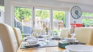 A dining area with a table set for a meal, chairs, a clock on the wall, and a view of greenery outside. - Stylish 3BR Home | Hot Tub | Near NEC, HS2 & JLR (West Midlands)