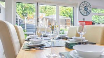 A dining area with a table set for a meal, chairs, a clock on the wall, and a view of greenery outside.