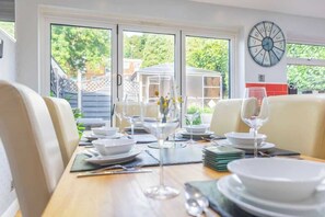 A dining area with a table set for a meal, chairs, a clock on the wall, and a view of greenery outside.