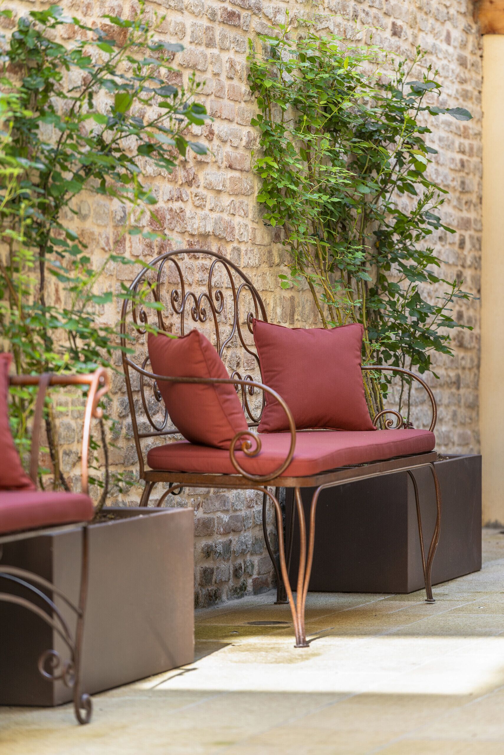 A wrought iron chair with red cushions against a brick wall, accompanied by a planter box.