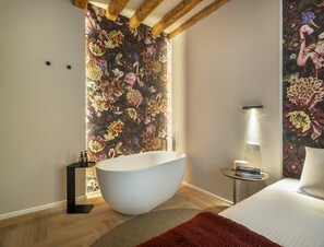 A modern bathroom with a freestanding bathtub, a round bedside table with a lamp, and a bed with a red rug. - Sarti Palace - Rose Apartment R&R (Venice)
