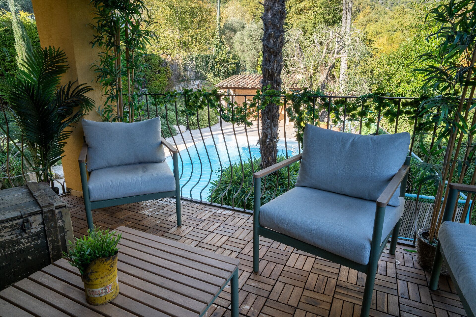 Eine Holzterrasse mit zwei grauen Sitzstühlen, einem kleinen Tisch mit einer Pflanze und einem Pool im Hintergrund.