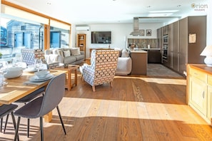 A modern interior with a dining area, sofa, kitchen, and a view of a wooden building outside. - Waters Edge 16, Heron Lodge - Lakeside Holiday Home, Hot Tub, Sleeps 8 (Cirencester)