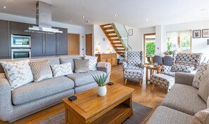 A modern living room with a grey sofa, patterned cushions, a wooden coffee table, and a staircase leading to an upper level. - Waters Edge 16, Heron Lodge - Lakeside Holiday Home, Hot Tub, Sleeps 8 (Cirencester)