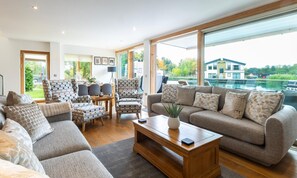 A spacious living room with a large window, a fireplace, and a view of a lake. - Waters Edge 16, Heron Lodge - Lakeside Holiday Home, Hot Tub, Sleeps 8 (Cirencester)