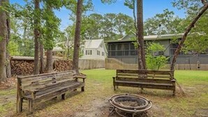 Un banco de madera en un patio con un brasero, rodeado de árboles y una casa al fondo.