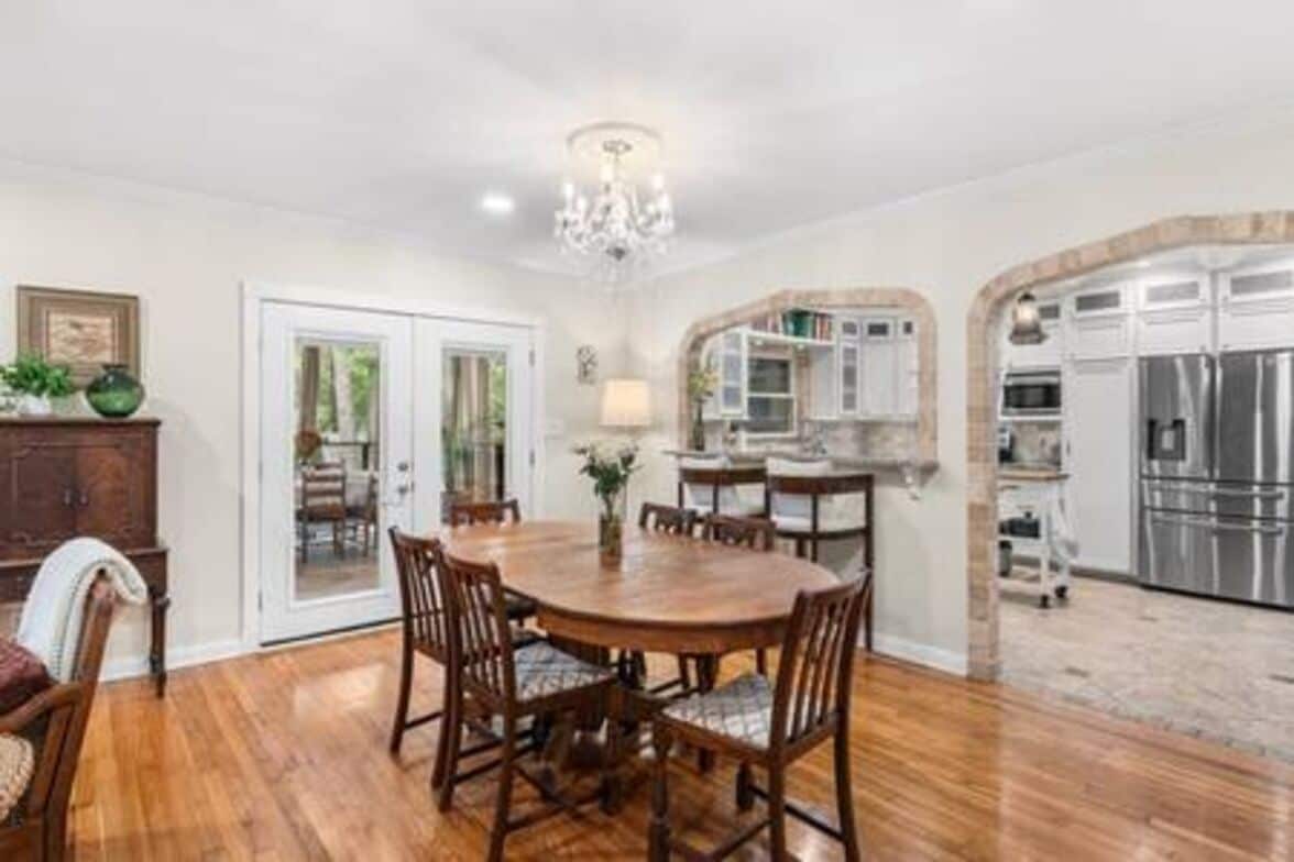 A dining area with a wooden table and chairs, a chandelier, and a kitchen with stainless steel appliances.