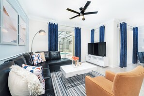 Ein modernes Wohnzimmer mit einem schwarzen Ledersofa, einem weißen Couchtisch und einem Fernseher auf einem weißen Schrank. Blaue Vorhänge sind vorhanden, und ein Deckenventilator ist sichtbar.