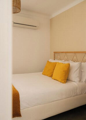 Cama con ropa blanca y dos almohadas amarillas, cabecera de madera y tapiz tejido en la pared.