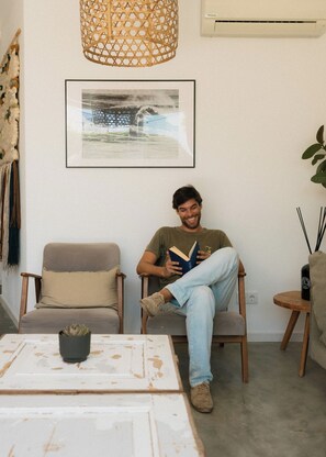 A person sitting on a chair reading a book in a room with a framed picture on the wall, a hanging light fixture, and a small table with a plant. - Outsite Ericeira Boavista (Mafra)