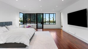 Ein modernes Schlafzimmer mit einem großen Bett, einem an der Wand montierten Fernseher und Blick auf Palmen durch große Fenster.