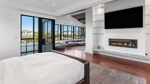 Ein modernes Schlafzimmer mit einem großen Bett, einem Kamin, einem Fernseher und einem Balkon mit Blick auf Palmen.