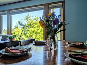 Ein Esstisch mit Tellern, einer Blumenvase und Blick auf Bäume durch große Fenster.