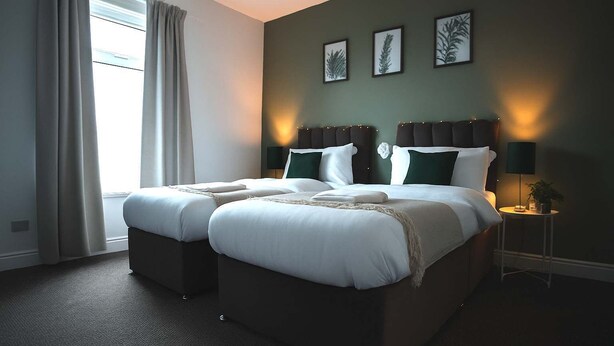 Ein Doppelbett mit Kopfteil, zwei Nachttische, ein Fenster mit Vorhängen und ein an der Wand befestigtes Kunstwerk.