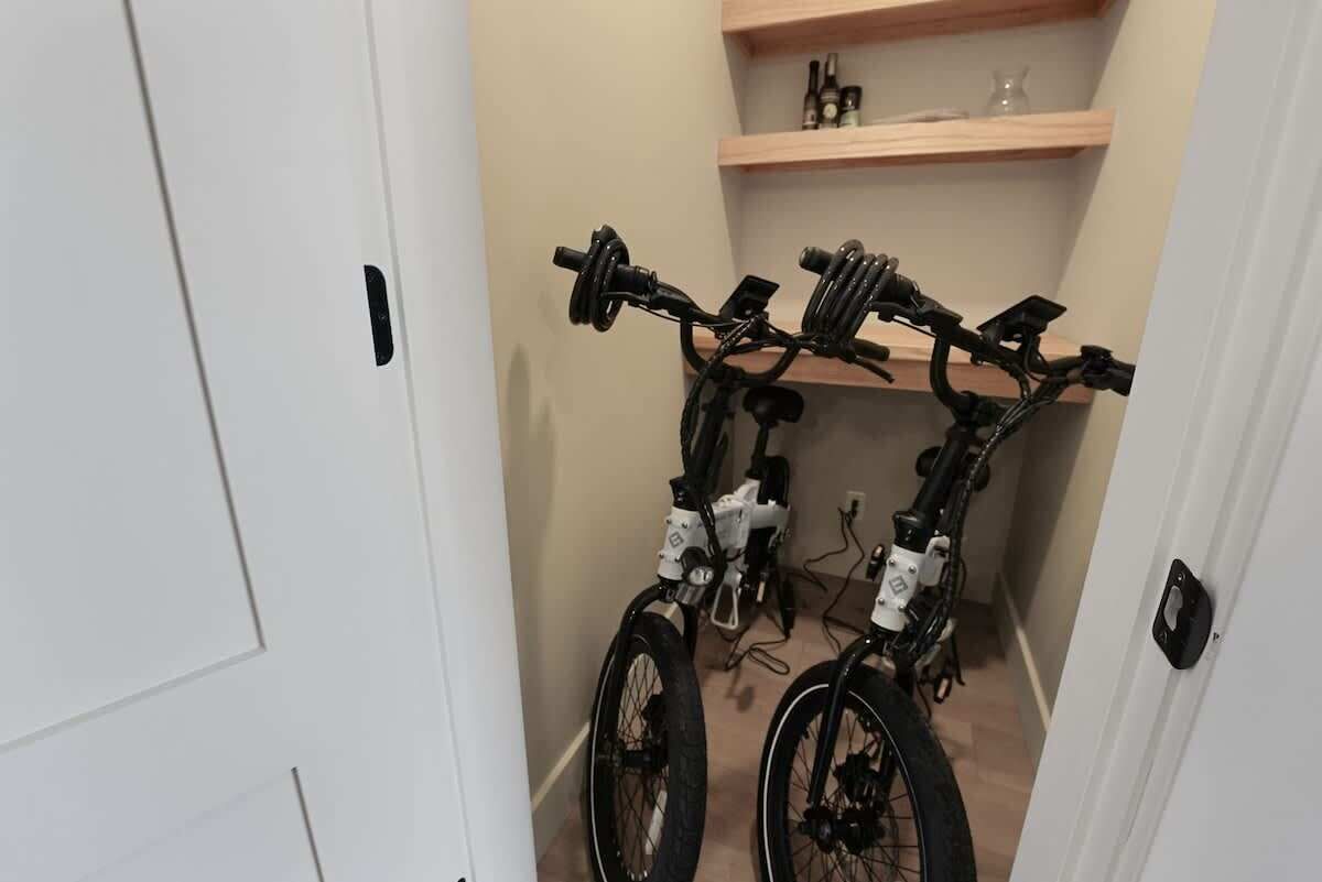 Een smalle gang met twee fietsen tegen de muur, een witte deur links en een houten plank met flessen en een vaas op de achtergrond.