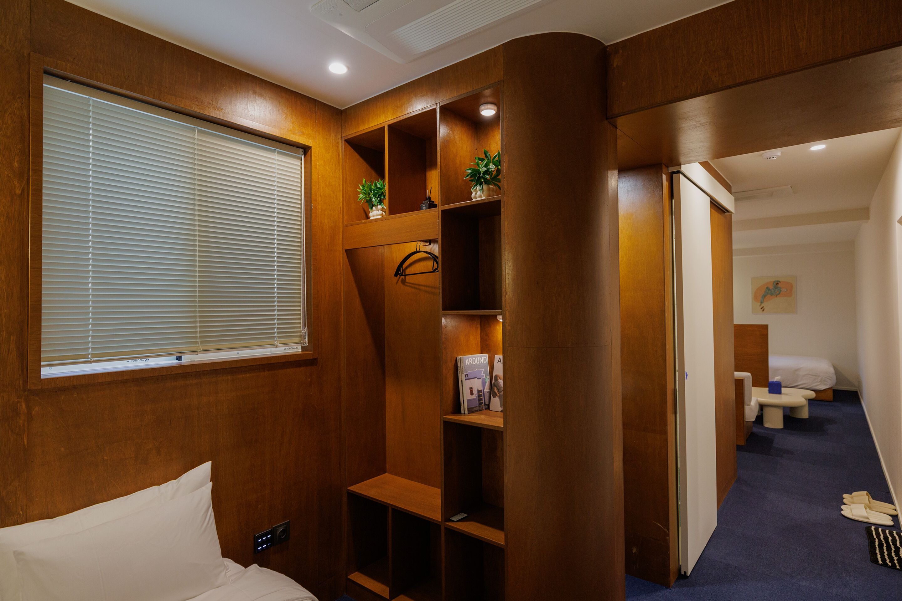 A hotel room with a wooden panel wall, a bed, a window with blinds, a shelf with plants, and a door leading to another room.