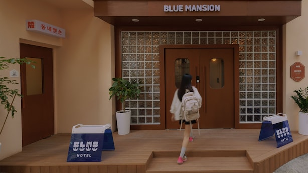 Entrada de hotel con un cartel que dice 'BLUE MANSION', una escalera de madera y dos plantas en macetas.