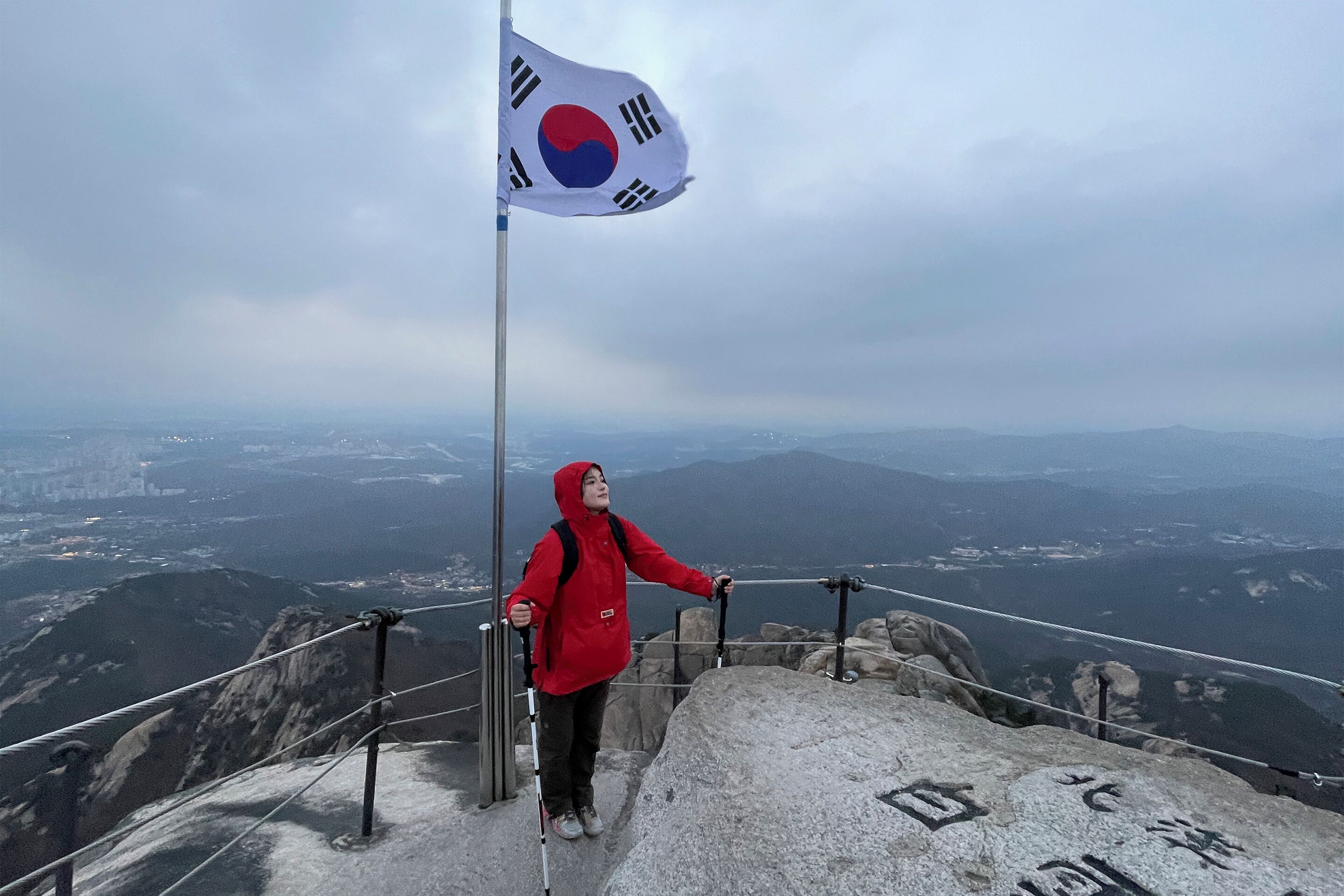 Une personne se tient au sommet d’une montagne, un drapeau à la main, et contemple un vaste paysage.
