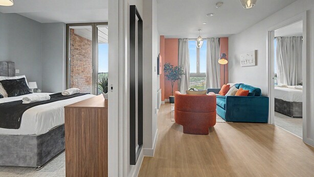 A modern hotel room with a bed, a seating area with a sofa and armchair, a potted plant, and a view of the city. - GuestReady - Chic 2-Bed Apartment with Balcony (Manchester)