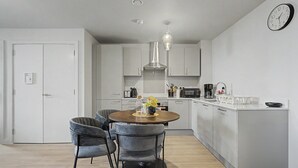 A modern kitchen with a dining area, featuring a round table, grey upholstered chairs, a hanging light fixture, and a clock on the wall. - GuestReady - Chic 2-Bed Apartment with Balcony (Manchester)