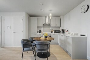 A modern kitchen with a dining area, featuring a round table, grey upholstered chairs, a hanging light fixture, and a clock on the wall. - GuestReady - Chic 2-Bed Apartment with Balcony (Manchester)