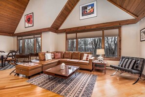 A spacious living room with a large sectional sofa, a coffee table, and a fireplace. There are large windows offering a view of trees outside.