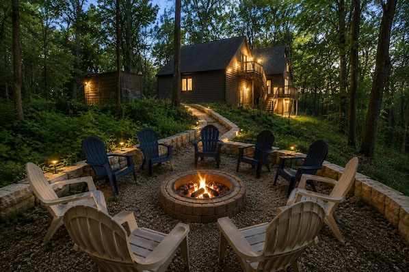 A fire pit surrounded by Adirondack chairs in a forest setting with a cabin in the background.