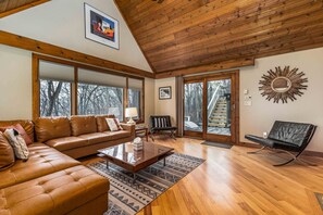 A spacious living room with a wooden ceiling, large windows, a sofa, a coffee table, and a chair.